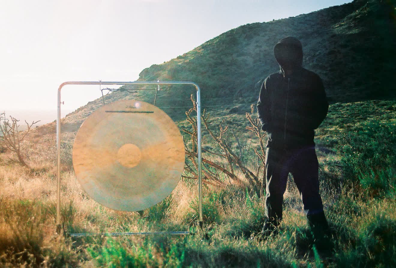 Gong Slayer standing beside a gong in dry grass below a hill